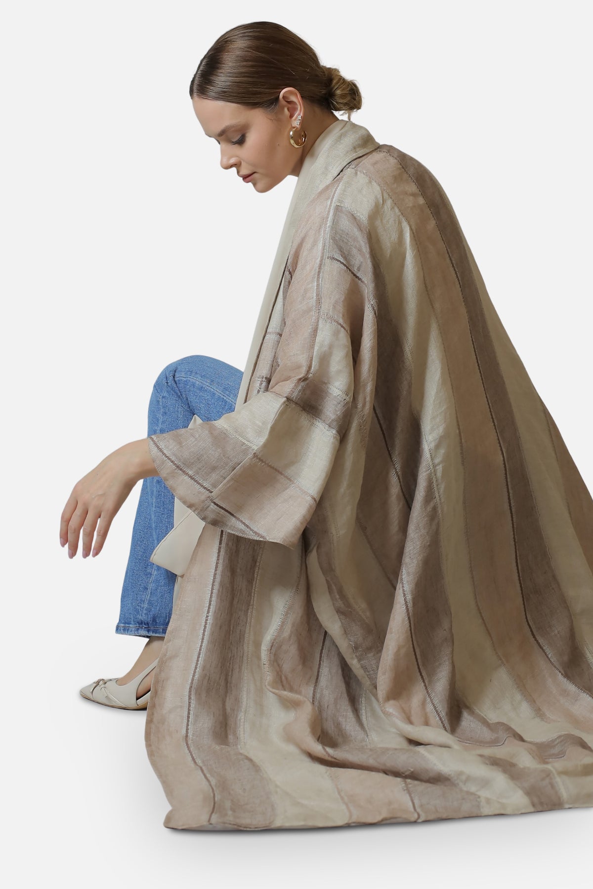 Seated side view of the neutral striped linen abaya showing the fabric drape and hem.