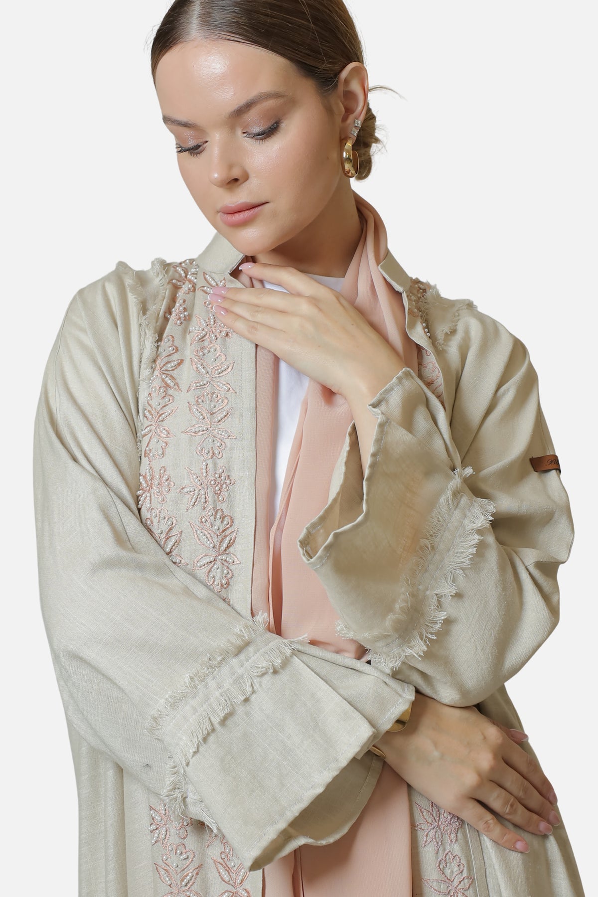 Beige linen abaya with pearl-embellished embroidery, worn with a blush shaila and one hand raised to the neckline.